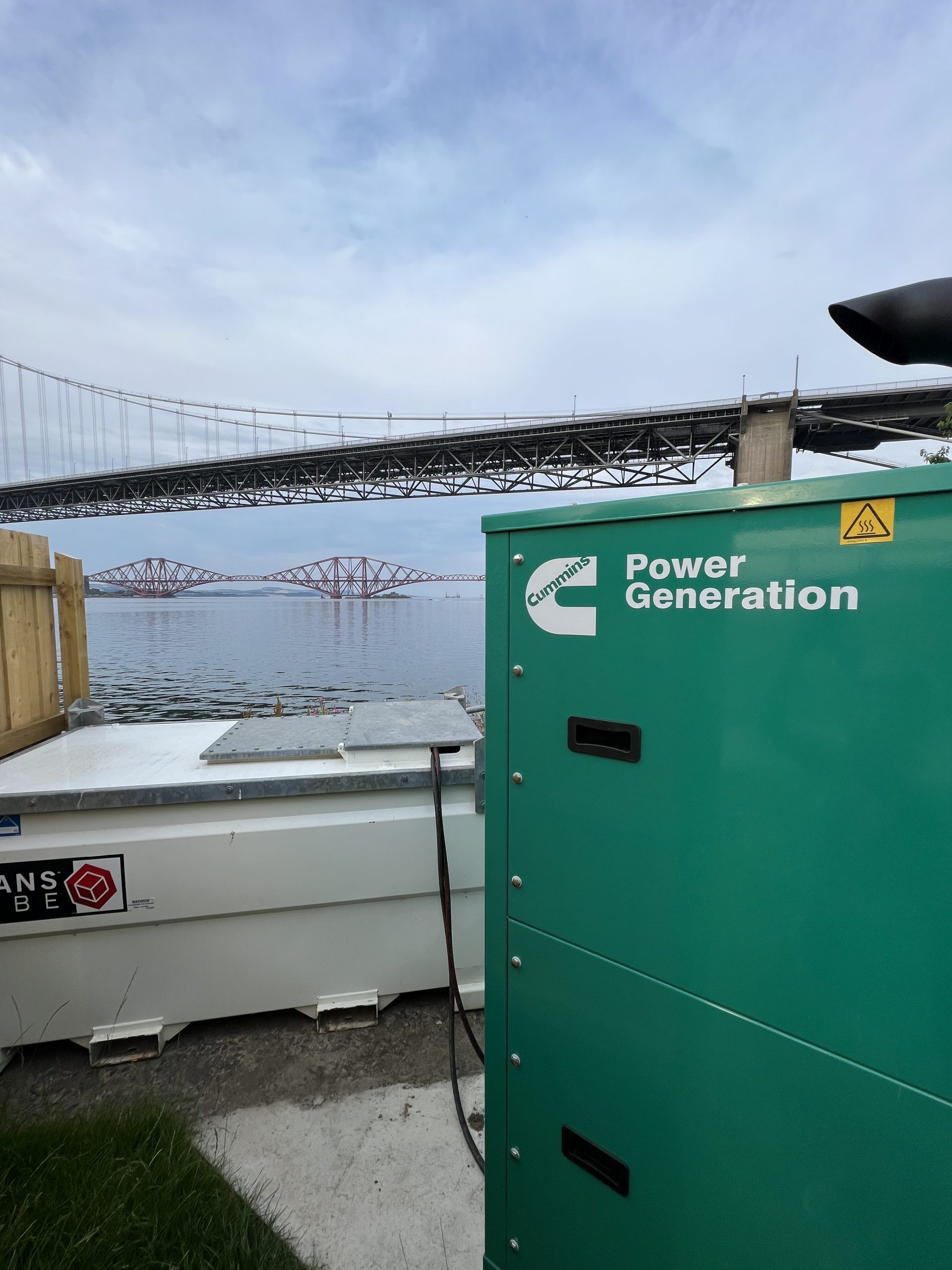 A green Cummins Generator connected to an external fuel tank against a backdrop of the forth bridges. This is to show some of the products that we have in our rental range.